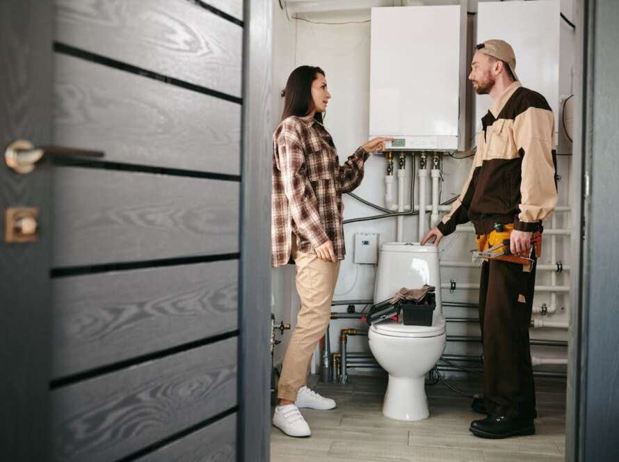 Plumber speaking to a woman and inspecting a toilet and pipes.