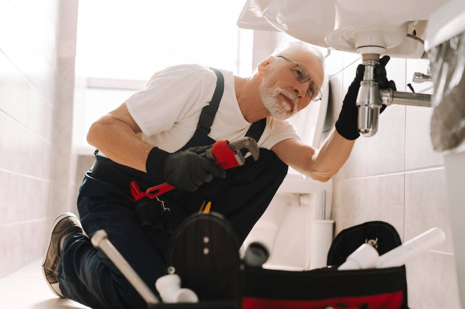 A plumber repairs a sink pipe with an adjustable wrench under the kitchen basin.