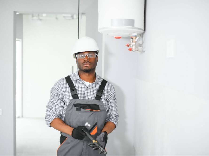 A man wearing a yellow hard hat checks a water heater in a commercial space with a tool.