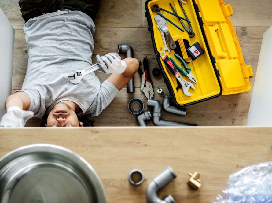 A plumber lying on his back with an open toolbox beside him, reaching up to perform plumbing maintenance on a kitchen sink with a crescent wrench.
