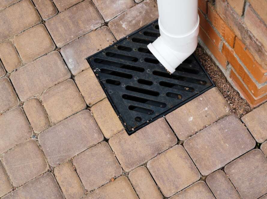 A storm drain grate installed on paved ground with a downspout connected for proper drainage.