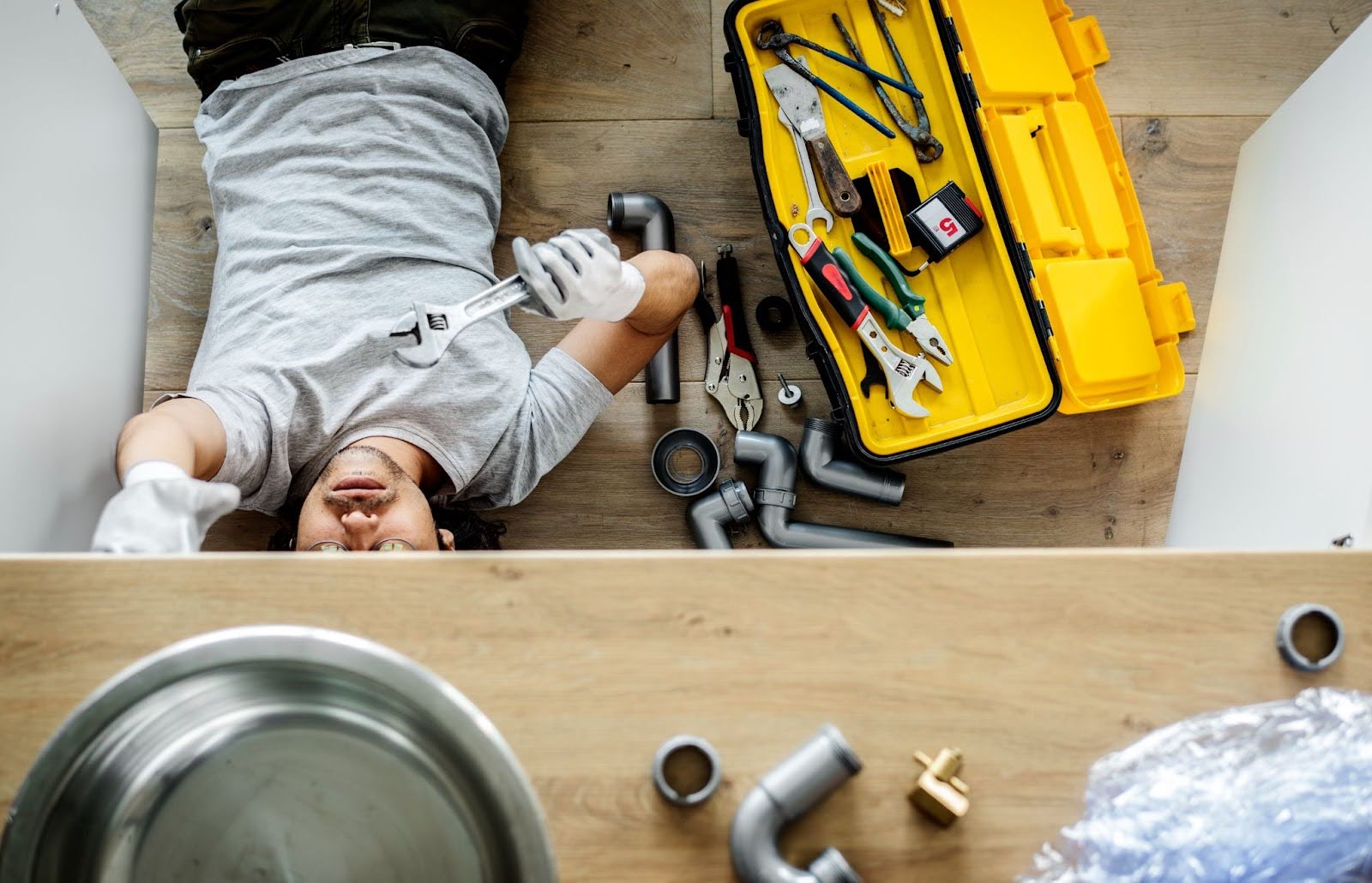A plumber lying on his back with an open toolbox beside him, reaching up to perform plumbing maintenance on a kitchen sink with a crescent wrench.