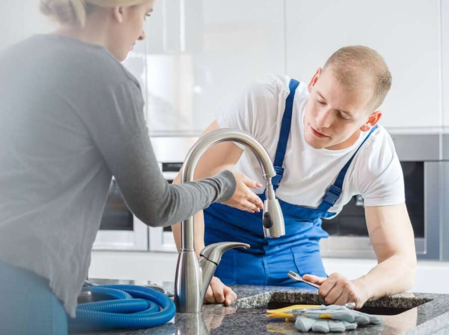 A homeowner shows the leaky kitchen faucet to the plumber in blue overalls.