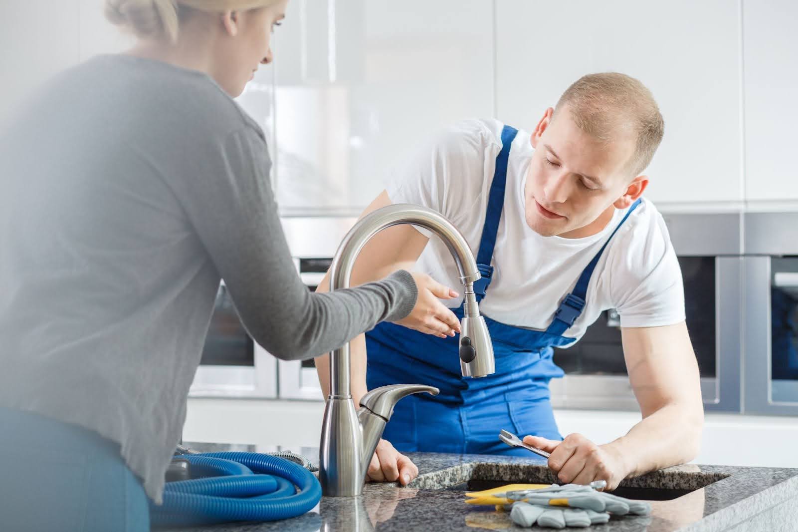 A homeowner shows the leaky kitchen faucet to the plumber in blue overalls.