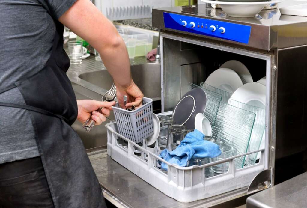 Restaurant kitchen staff member unloading clean dishes from a commercial dishwasher.