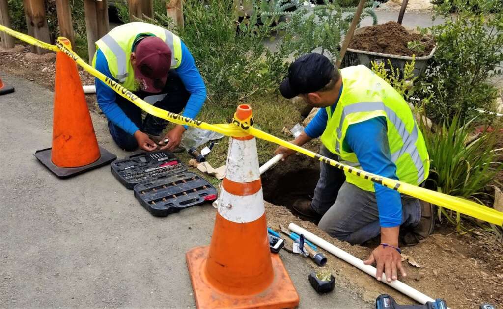 Plumbers performing a sewer line cleanout with yellow caution tape and orange safety cones marking the work area for public safety.