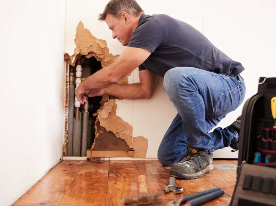 Emergency plumber repairing a burst pipe inside a cracked interior wall.