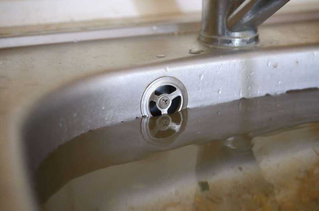 Close-up of a stainless steel sink drain filled with standing water caused by a clogged sewer line.