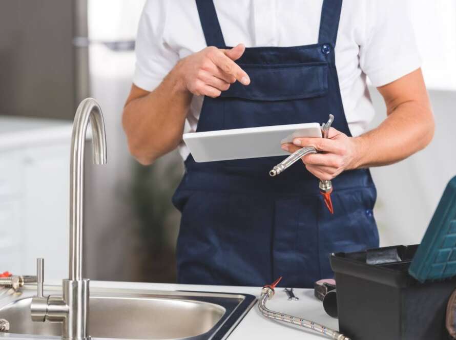 Commercial plumber reviewing installation details on a tablet before installing a garbage disposal unit in a restaurant kitchen.