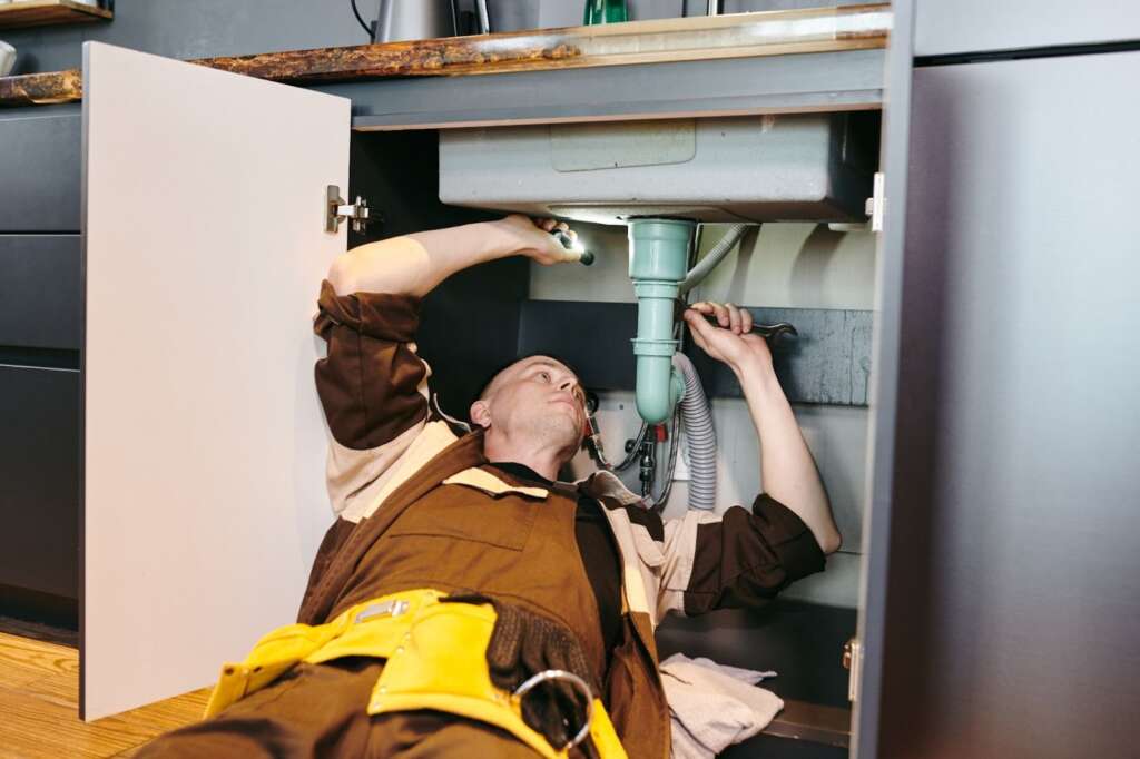 A plumber installing garbage disposal piping under a commercial kitchen sink using a wrench.