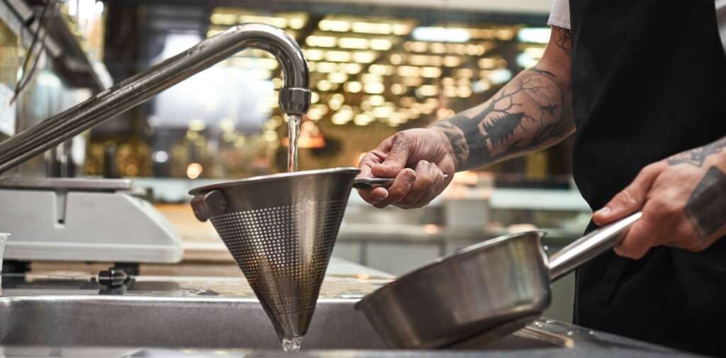 Close-up photo of a chef’s tattooed hands holding cooked pasta in a colander under running water